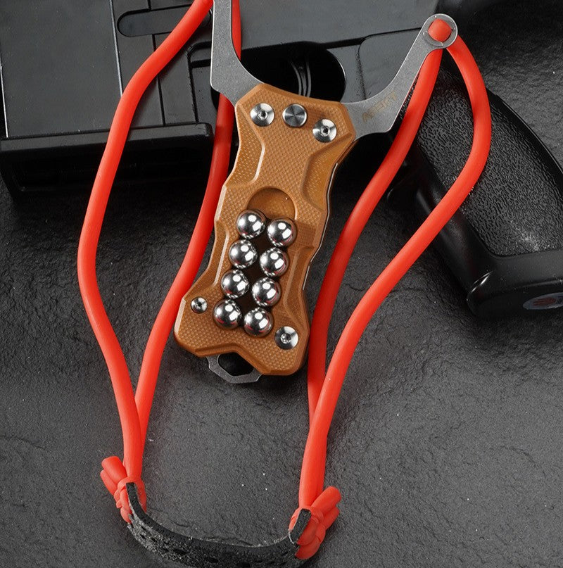 A tan G10 handle EDC slingshot with a red high-elastic band and steel ball storage, displayed with the band hung loosely on a dark tactical firearm background.