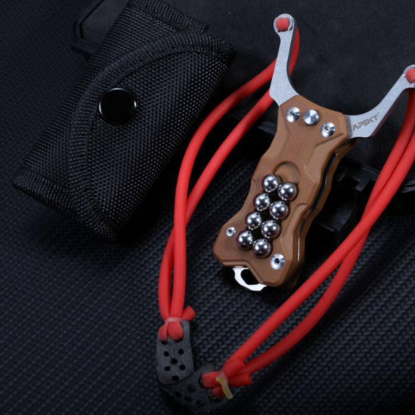 A tan G10 handle EDC slingshot with a red band and steel balls, displayed next to a black nylon sheath on a dark textured surface.
