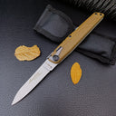 An open tan-handled folding knife displayed with its black nylon carrying pouch and dried yellow leaves on a leather surface.