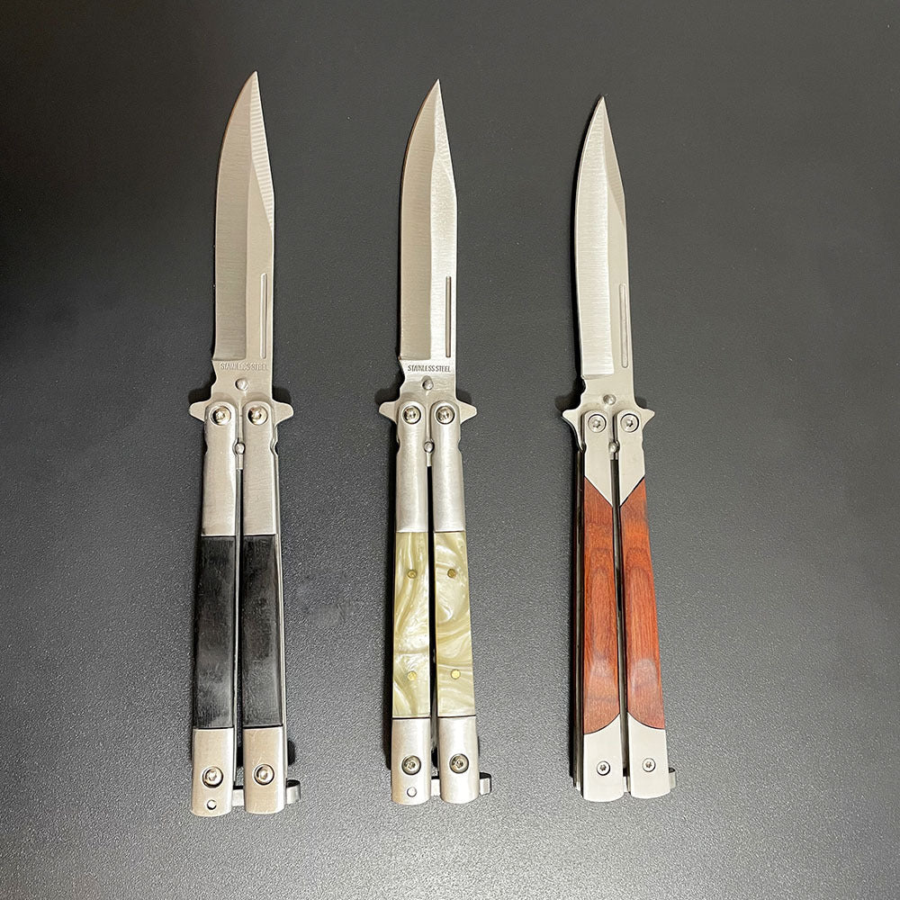 Three open balisong butterfly knives with silver blades and different handle materials: wood, pearl, and black, arranged vertically on a dark background.