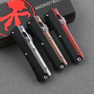 Three closed out-the-front knives with black, bronze, and red handles resting on a black box with a red octopus logo, displayed on a dark textured surface.