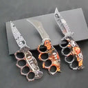 Three open folding knuckle duster knives with unique custom graphics including a damascus-style etched blade, a BROWNING cutout blade, and an orange skull handle with a serrated blade, displayed on a dark surface.