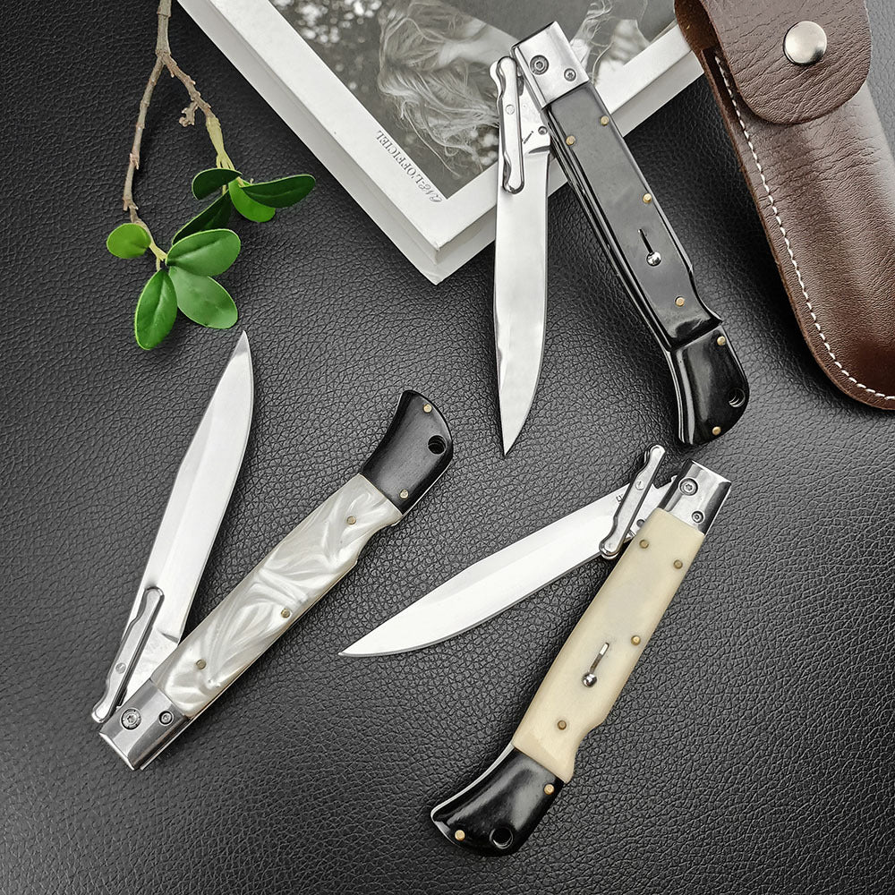 Three Italian stiletto folding knives featuring black, pearl white, and cream bone handles, displayed on a dark leather surface alongside a book, green leaves, and a brown leather sheath.