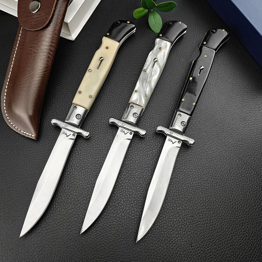 Three open Italian stiletto folding knives with polished silver blades and crossguards. The handles are black, black-and-white pearl/marbled, and cream bone. They are displayed on a dark textured leather surface next to a brown leather sheath and green leaves.
