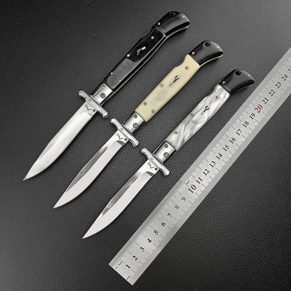 Three open Italian stiletto folding knives with black, cream bone, and white pearl handles, displayed diagonally next to a metal ruler for size reference on a dark leather surface.