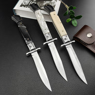 Three open Italian stiletto folding knives with polished silver blades and crossguards, arranged diagonally on a dark textured leather surface. The handles from left to right are black, pearlescent white, and cream bone. The setup is styled with a book, a sprig of green leaves, and a brown leather sheath.