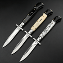 Three open Italian stiletto folding knives with silver blades and crossguards, featuring black, creamy white, and black-and-white marbled handles, arranged diagonally on a dark background.