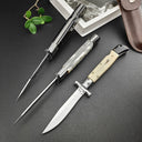 Three open Italian stiletto folding knives with silver blades displayed on a dark leather surface. The handles feature black, white pearlescent, and cream bone finishes. The composition includes a brown leather sheath, a book, and green leaves in the background.
