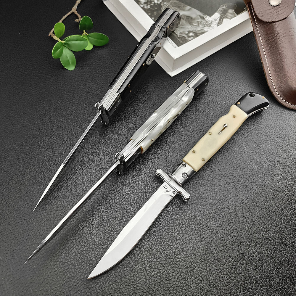 Three open Italian stiletto folding knives with silver blades displayed on a dark leather surface. The handles feature black, white pearlescent, and cream bone finishes. The composition includes a brown leather sheath, a book, and green leaves in the background.