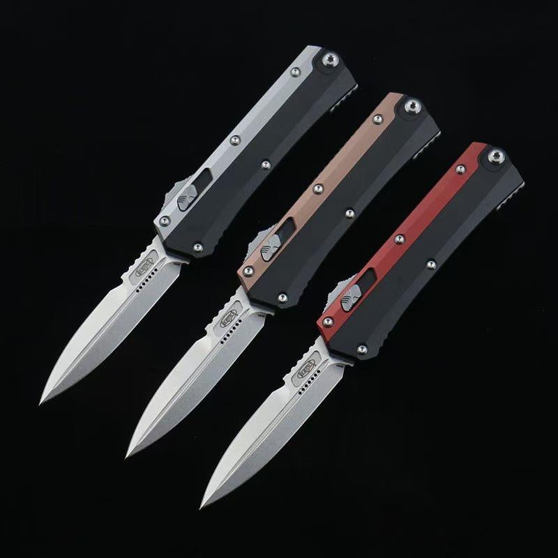 Three open out-the-front tactical knives with silver blades and black, bronze, and red handle accents, displayed on a dark background.