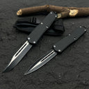 Two black OTF knives with silver blades and different blade shapes, displayed with a black nylon sheath on a rough textured surface with wood.
