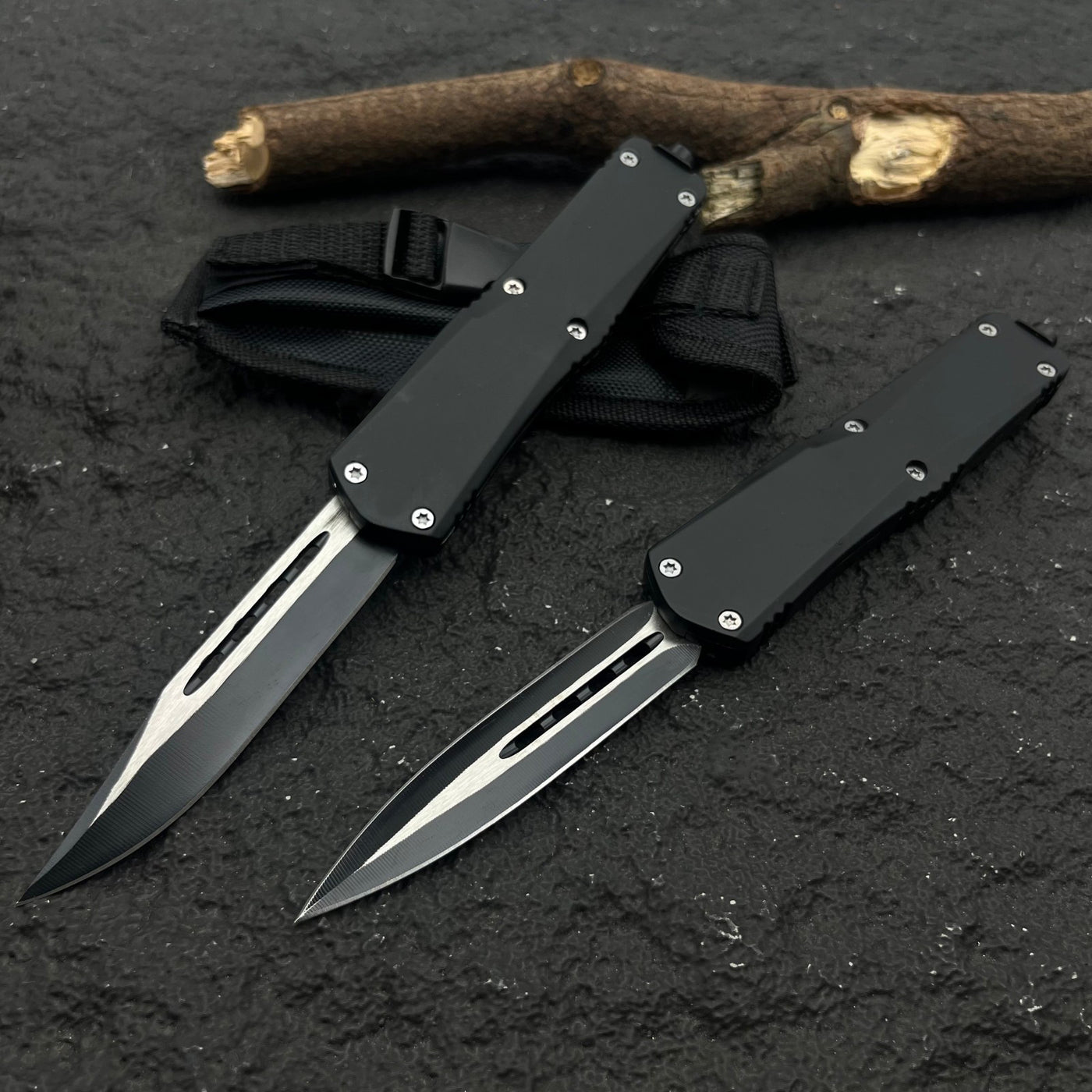 Two black OTF knives with silver blades and different blade shapes, displayed with a black nylon sheath on a rough textured surface with wood.