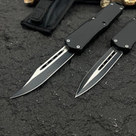 Two black OTF knives with silver blades and different blade shapes, displayed with a black nylon sheath on a rough textured surface with wood.