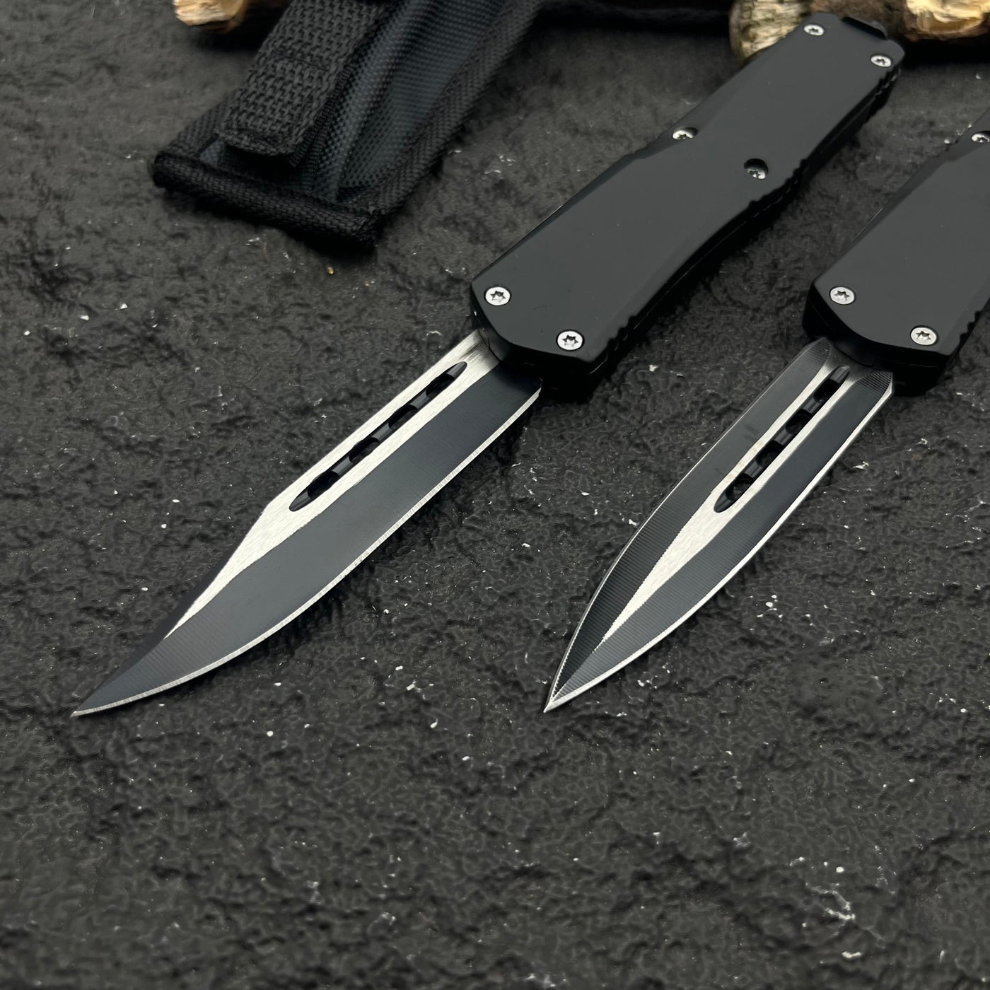 Two black OTF knives with silver blades and different blade shapes, displayed with a black nylon sheath on a rough textured surface with wood.