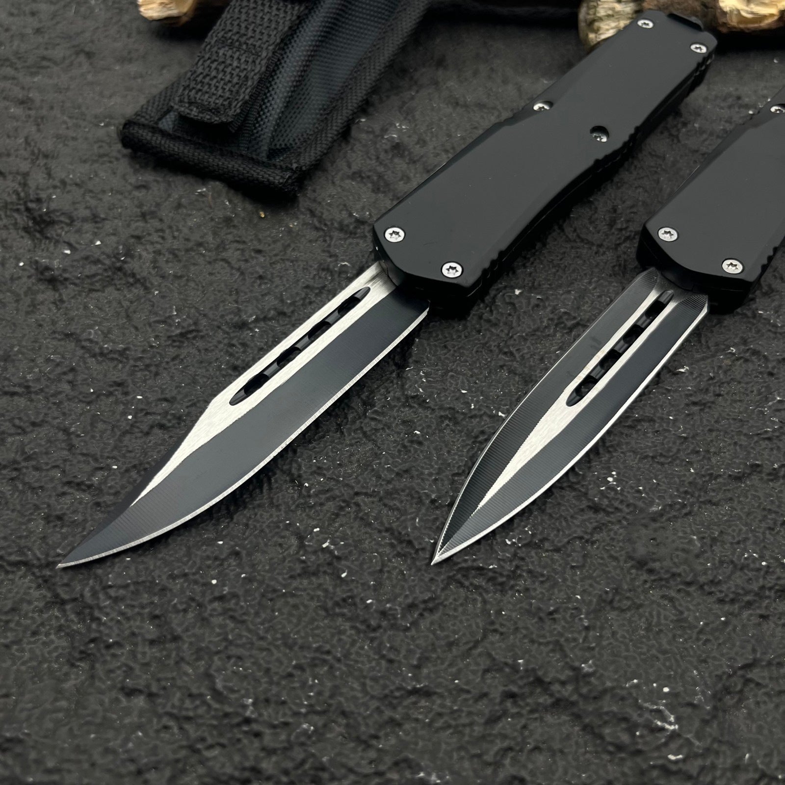 Two black OTF knives with silver blades and different blade shapes, displayed with a black nylon sheath on a rough textured surface with wood.