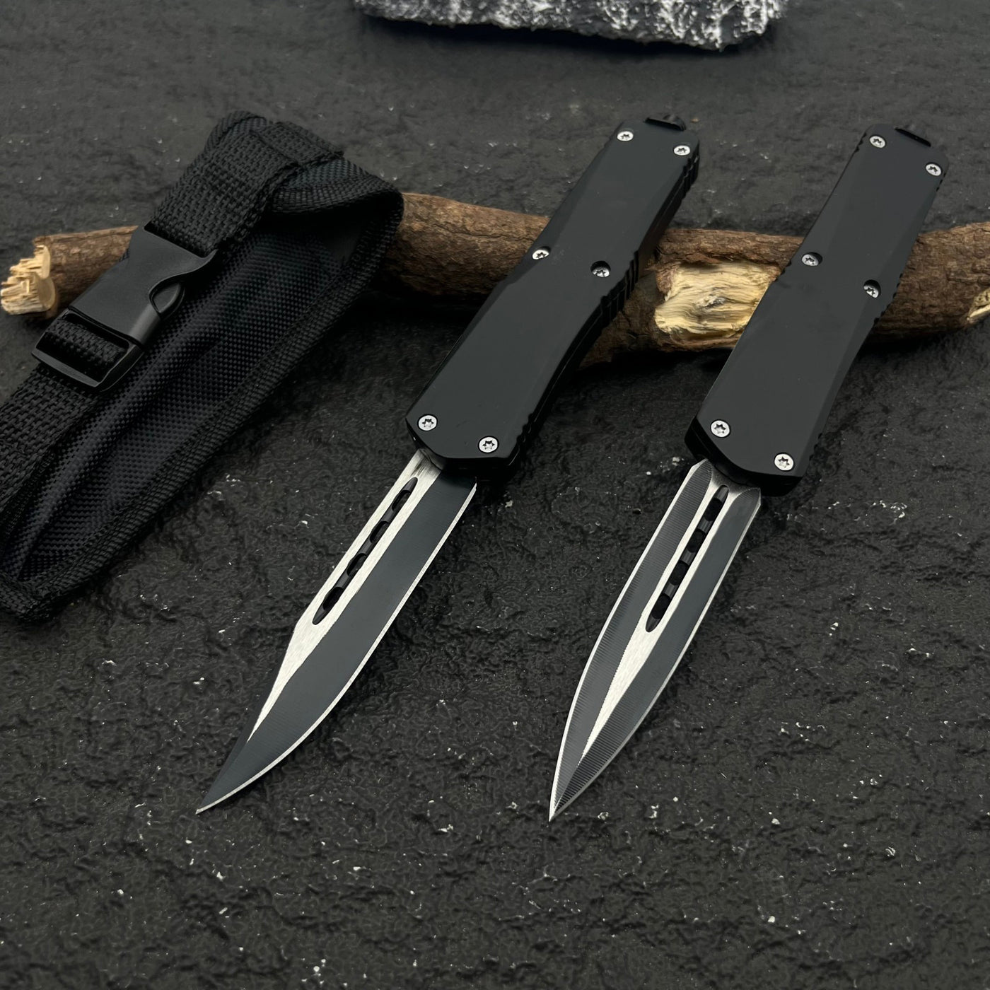 Two black OTF knives with silver blades and different blade shapes, displayed with a black nylon sheath on a rough textured surface with wood.