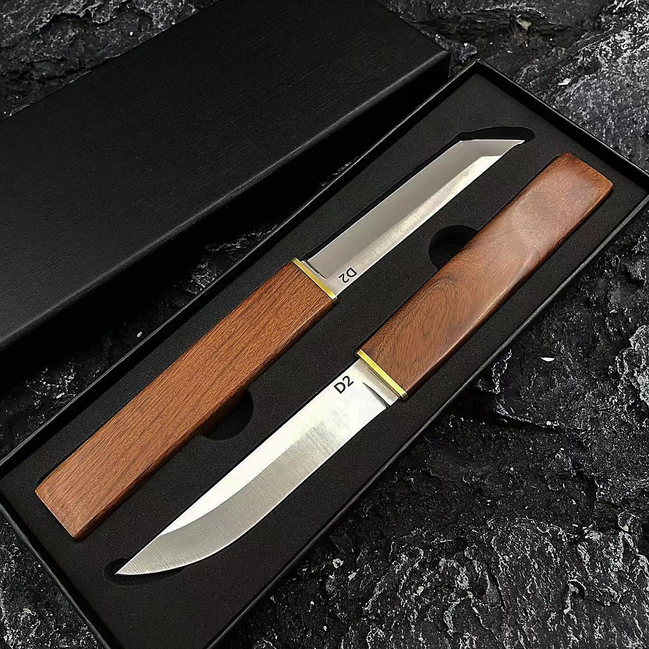 Two D2 steel fixed blade knives with wooden handles and brass accents displayed in a black box. One features a tanto blade, and the other features a curved blade, placed against a dark, rough rocky background.