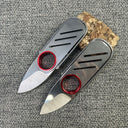 Two open EDC folding knives with silver bodies and red rings, displayed next to a small wood block on a grey textured surface.