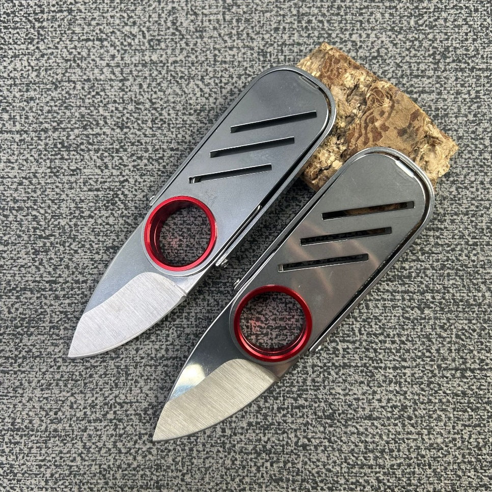 Two open EDC folding knives with silver bodies and red rings, displayed next to a small wood block on a grey textured surface.