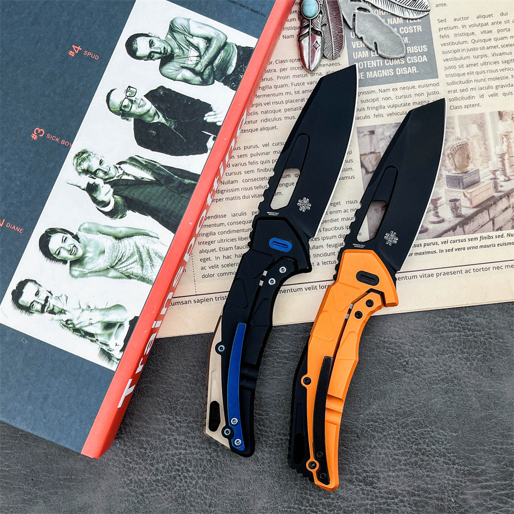 Two open folding knives with black blades and geometric handles. The left knife features a black and tan handle with a blue pocket clip and blue pivot, while the right knife has an orange handle with black accents. They are displayed on an open vintage magazine page with a feather pendant and a grey textured surface.