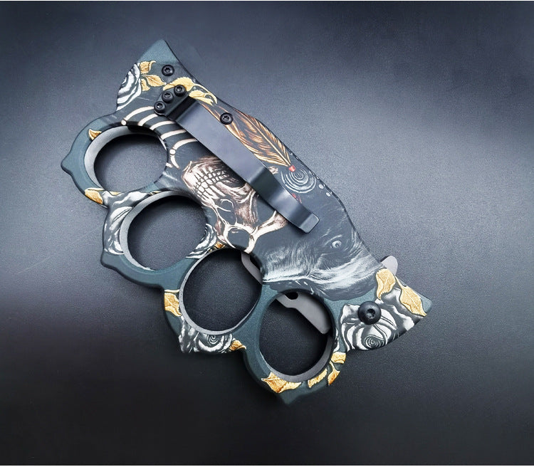 The back side of a closed knuckle duster folding knife, showing the black pocket clip and the continuation of the wolf and skull graphic on the handle.
