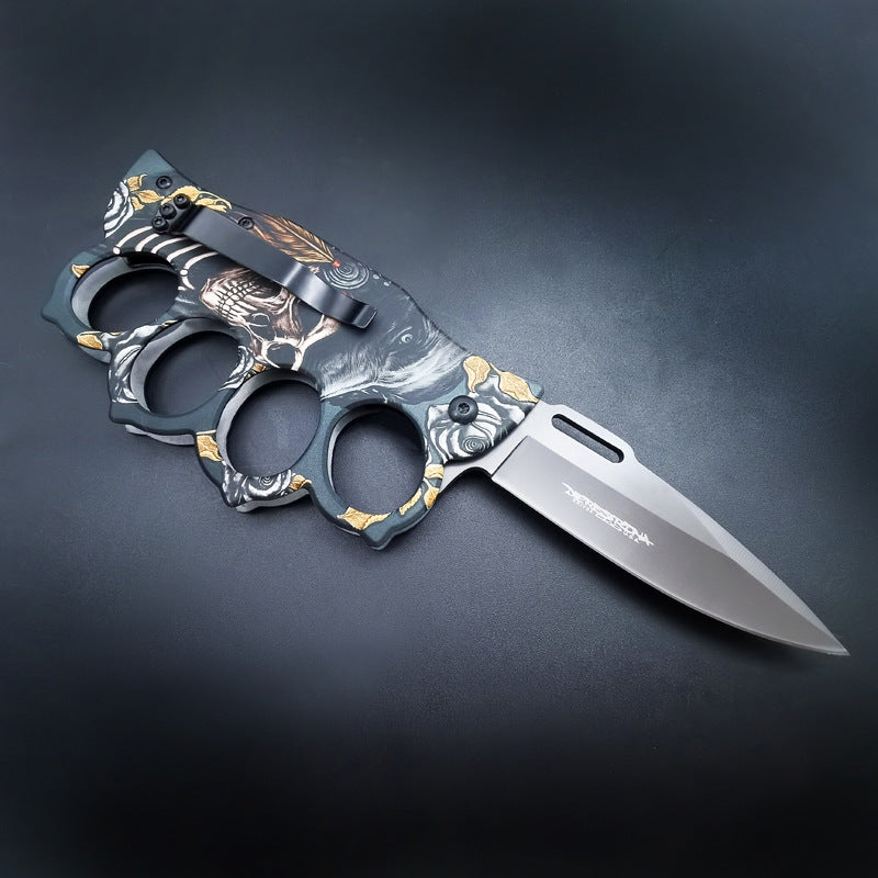 The back side of an open knuckle duster folding knife, highlighting the black pocket clip, textured handle, and the blade logo, on a dark background.