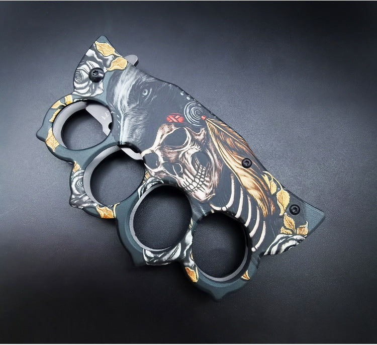 A closed folding knuckle duster knife showcasing the detailed wolf, skull, and gold rose graphic design on the handle, on a dark background.