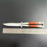 An open balisong butterfly knife with a wooden handle shown next to a metal ruler on a dark surface to display its length dimensions.