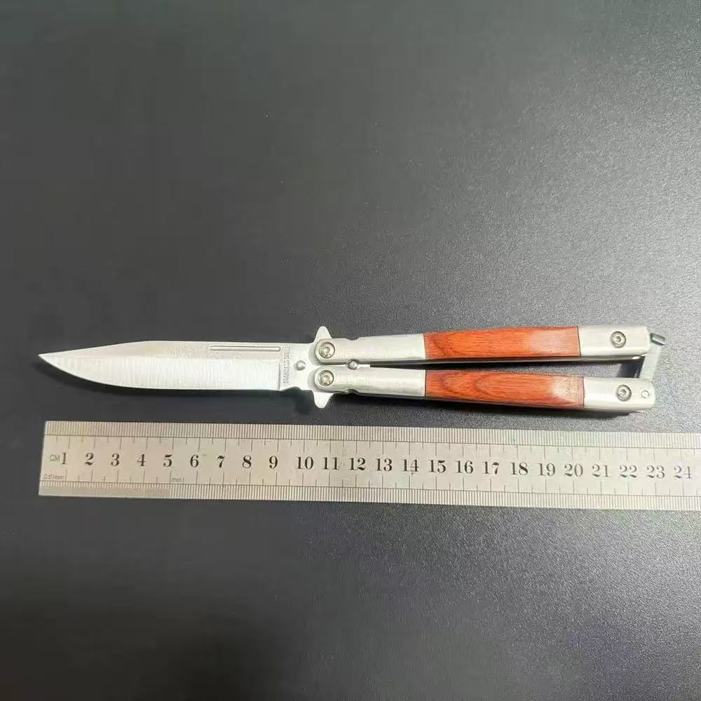 An open balisong butterfly knife with a wooden handle shown next to a metal ruler on a dark surface to display its length dimensions.