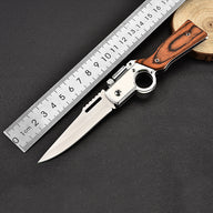 An open folding knife with a silver blade, wooden handle, and finger ring mechanism, shown next to a metal ruler for size reference on a dark surface.