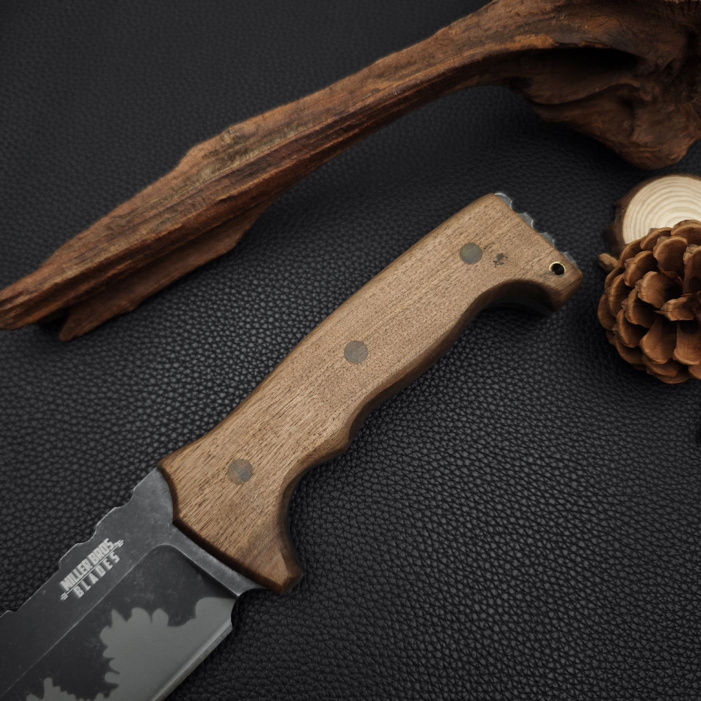 Close-up of a fixed blade knife's wooden handle with brass pins, displayed on a dark textured surface with pine cones and driftwood.