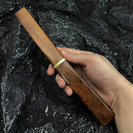 A hand holding a long, wooden knife sheath with a brass ring, displayed outdoors against a dark, rough rocky background.