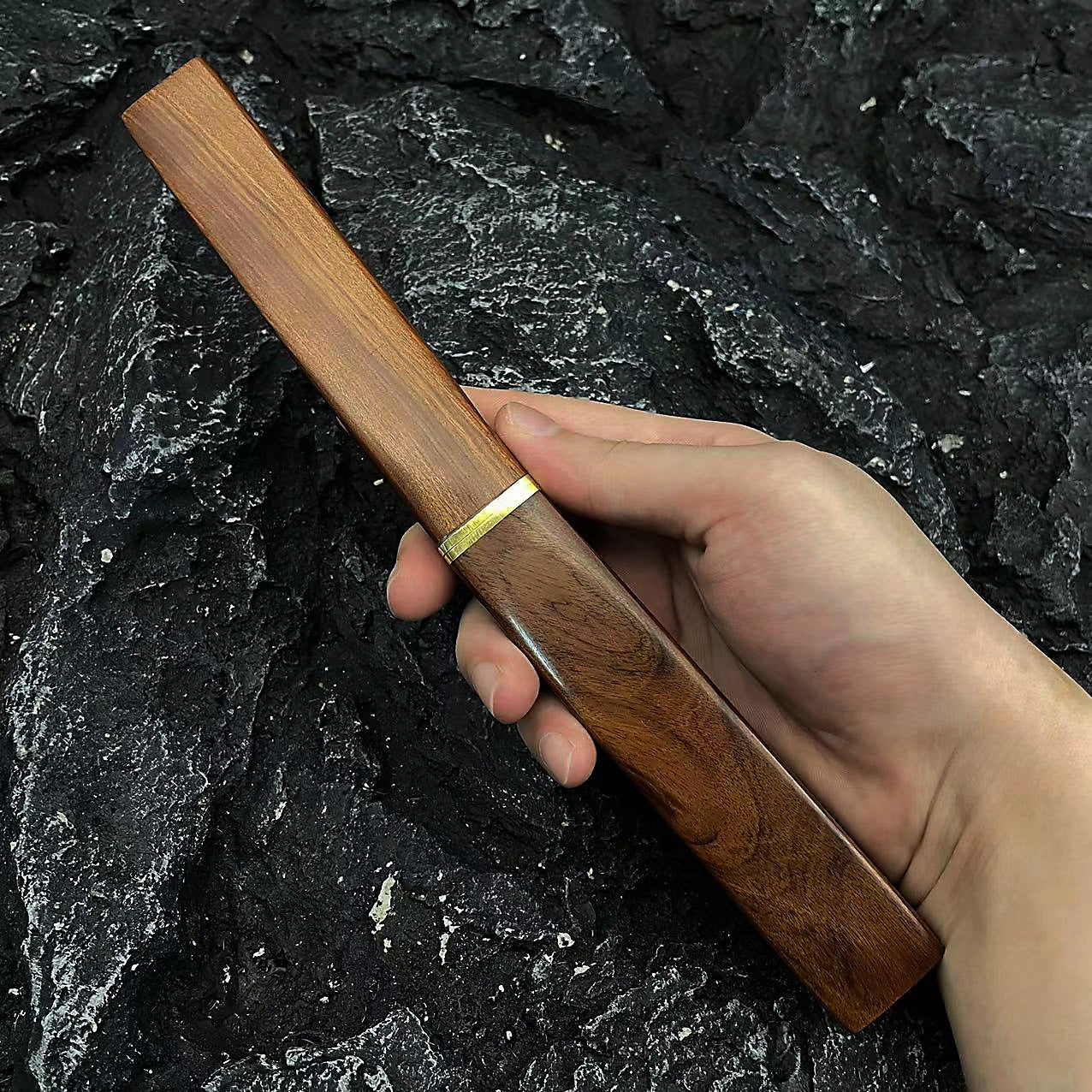 A hand holding a long, wooden knife sheath with a brass ring, displayed outdoors against a dark, rough rocky background.
