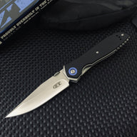 An open tactical folding knife with a black G10 handle, stonewashed silver blade, and blue pivot, displayed next to its black packaging box on a textured surface.