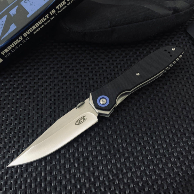 An open tactical folding knife with a black G10 handle, stonewashed silver blade, and blue pivot, displayed next to its black packaging box on a textured surface.