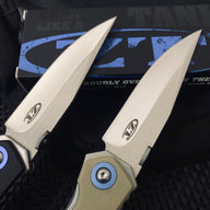 A close-up of the silver blades of two Zero Tolerance knives, showing the ZT logo and engraving, with a black packaging box in the background.