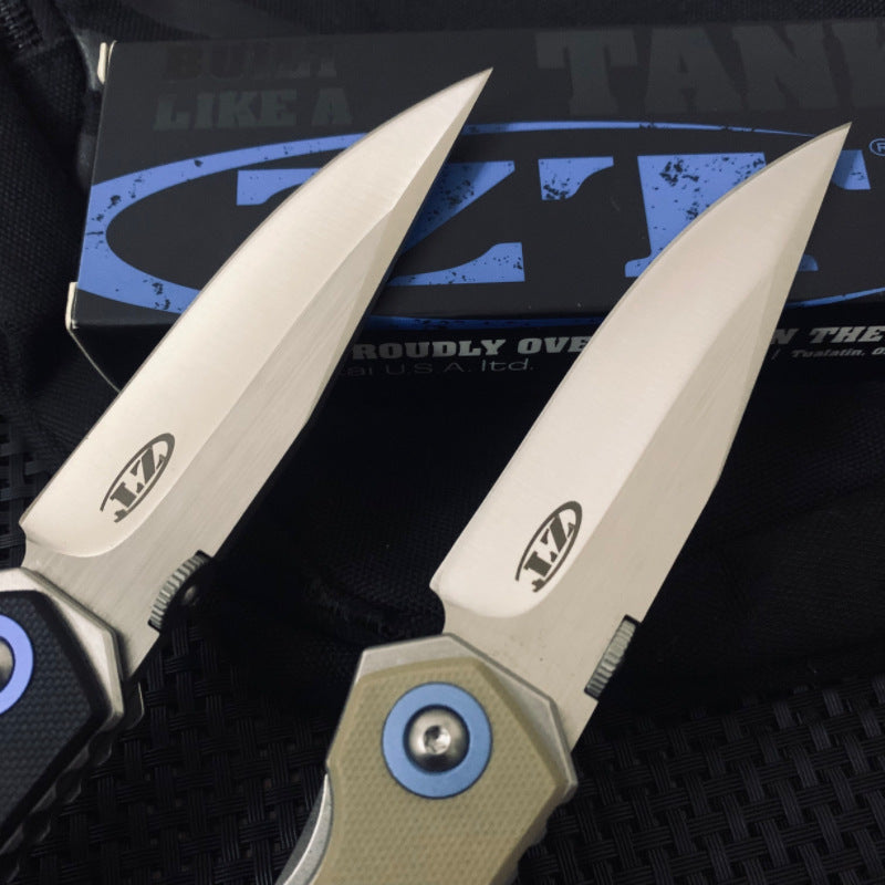 A close-up of the silver blades of two Zero Tolerance knives, showing the ZT logo and engraving, with a black packaging box in the background.