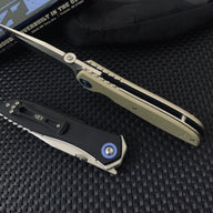 An open Zero Tolerance folding knife with a tan handle and silver blade, and a closed Zero Tolerance folding knife with a black handle and blue pivot screw, displayed on a textured black mat with a black box in the background.