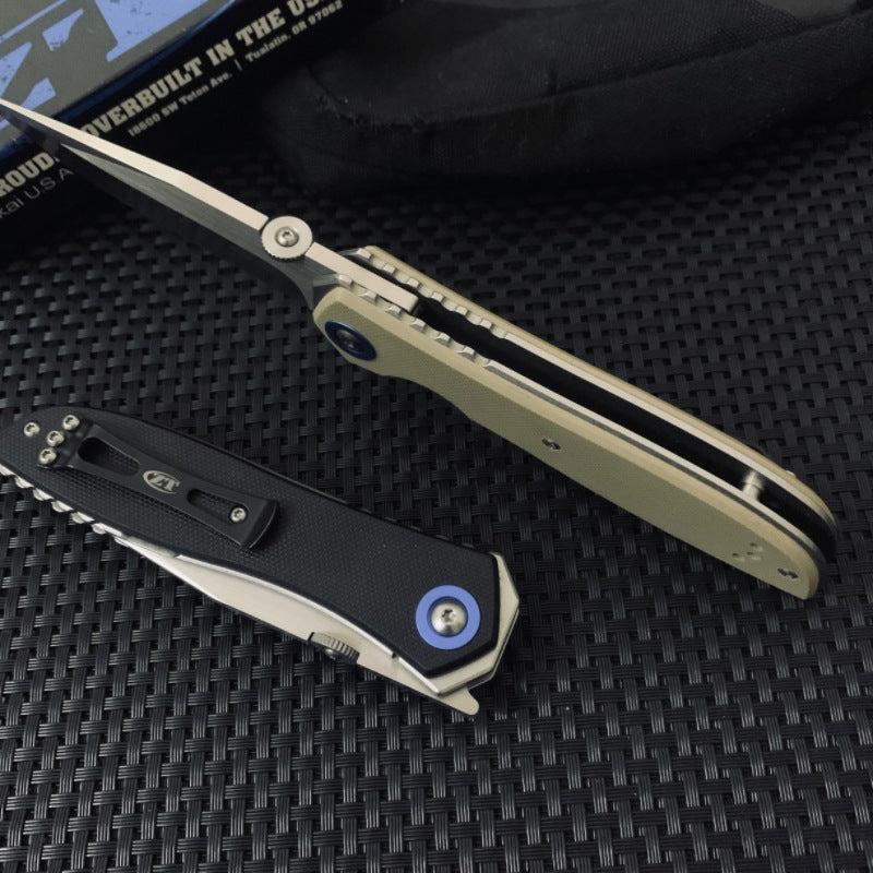 An open Zero Tolerance folding knife with a tan handle and silver blade, and a closed Zero Tolerance folding knife with a black handle and blue pivot screw, displayed on a textured black mat with a black box in the background.