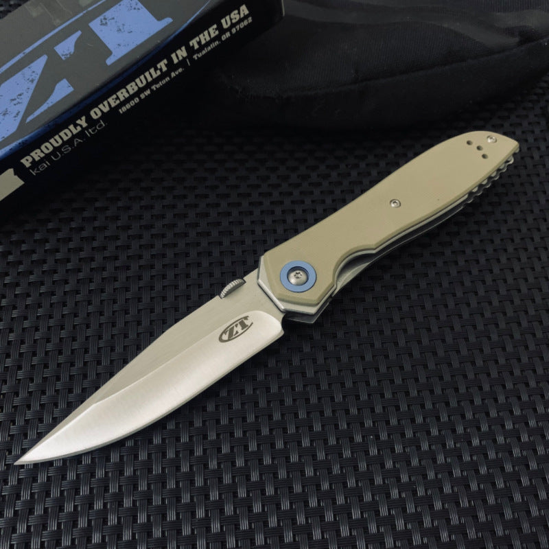 An open tactical folding knife with a tan G10 handle, stonewashed silver blade, and blue pivot, displayed next to its black packaging box on a textured surface.