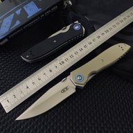 Two Zero Tolerance knives, one closed black and one open tan, displayed next to a metal ruler and a black box on a dark textured surface.
