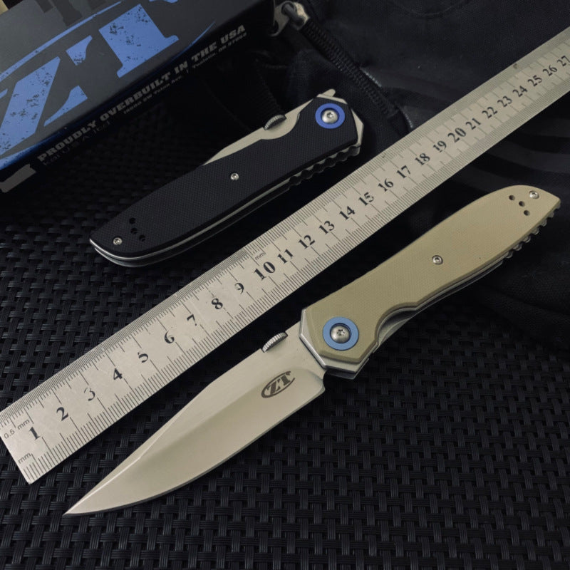 Two Zero Tolerance knives, one closed black and one open tan, displayed next to a metal ruler and a black box on a dark textured surface.