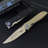 A closed black Zero Tolerance knife and an open tan Zero Tolerance knife, displayed on a dark textured surface with a box and pouch in the background.