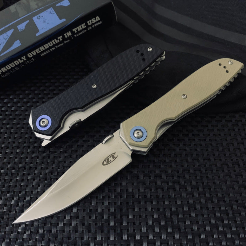 A closed black Zero Tolerance knife and an open tan Zero Tolerance knife, displayed on a dark textured surface with a box and pouch in the background.