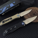 Two open Zero Tolerance knives, one tan and one black, resting on a wooden slice next to a box, showing the blade engraving on the tang.