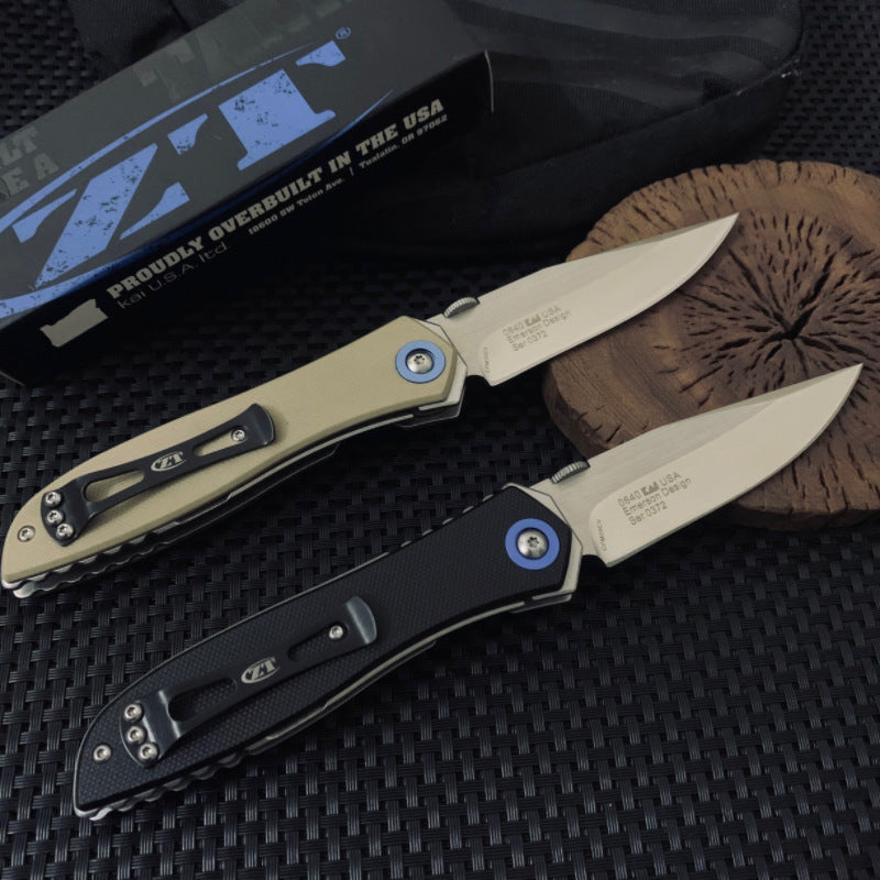 Two open Zero Tolerance knives, one tan and one black, resting on a wooden slice next to a box, showing the blade engraving on the tang.