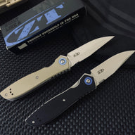 Two open Zero Tolerance folding knives with tan and black handles, displayed diagonally on a dark textured surface with a box in the background.