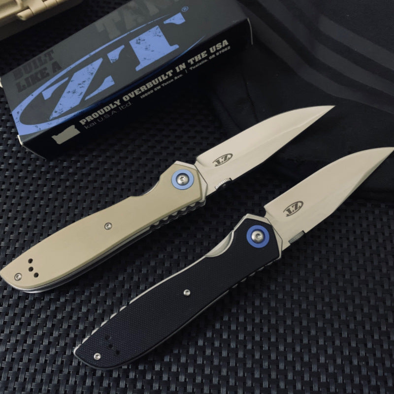 Two open Zero Tolerance folding knives with tan and black handles, displayed diagonally on a dark textured surface with a box in the background.