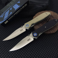 Two open Zero Tolerance knives, one with a tan handle and one with a black handle, resting on a wooden slice next to a black packaging box on a dark textured surface.
