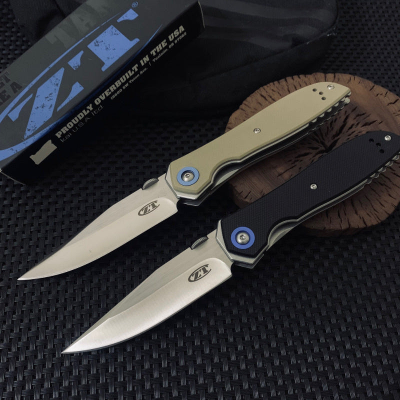Two open Zero Tolerance knives, one with a tan handle and one with a black handle, resting on a wooden slice next to a black packaging box on a dark textured surface.